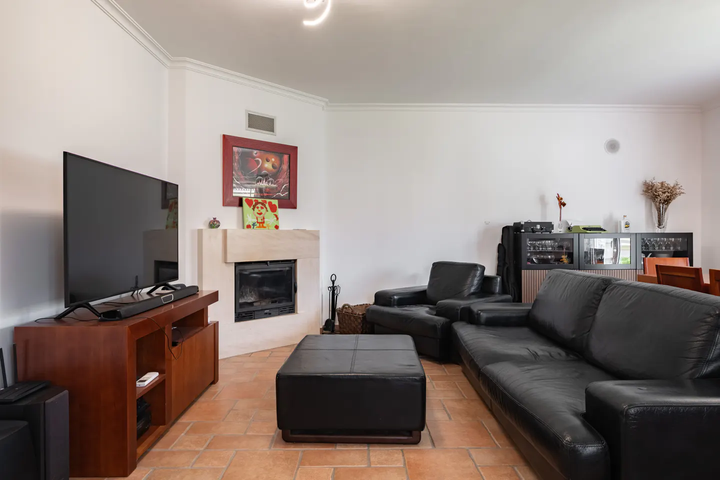 Living room with black leather sofa, chair, and ottoman. A TV sits on a wooden stand next to a fireplace. The floor is tiled.