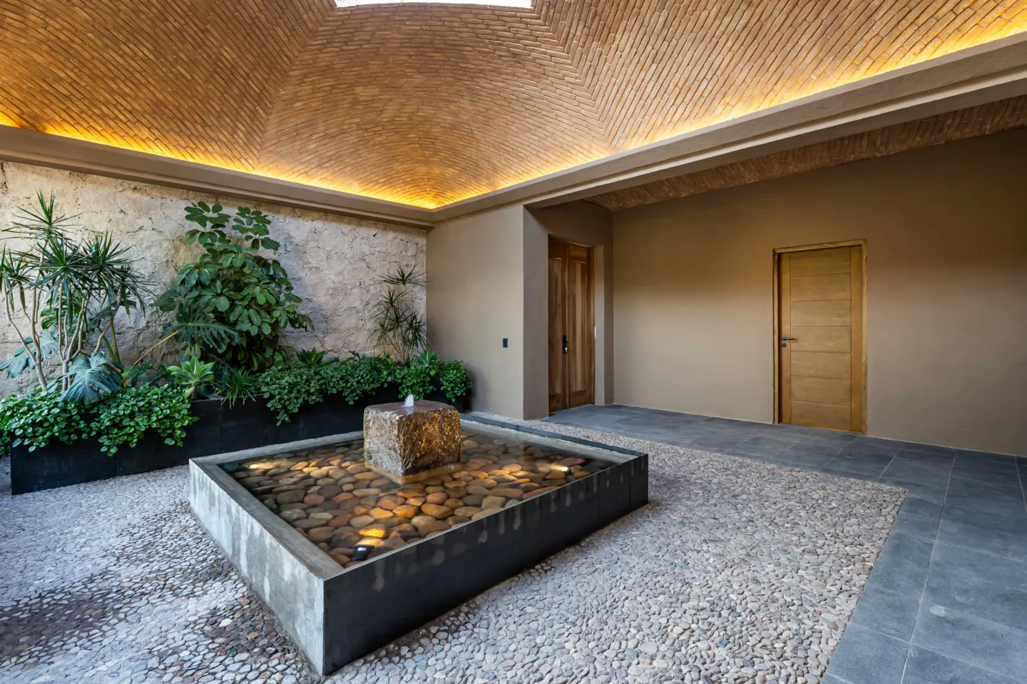 Indoor courtyard with stone fountain, pebble ground, and brick vaulted ceiling with skylight. Green plants line a stone wall.