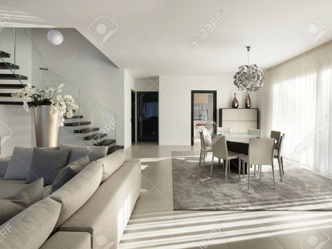 Bright, modern living room with a gray sofa, glass-railed stairs, and a dining table with white chairs. Sunlight streams through sheer curtains.