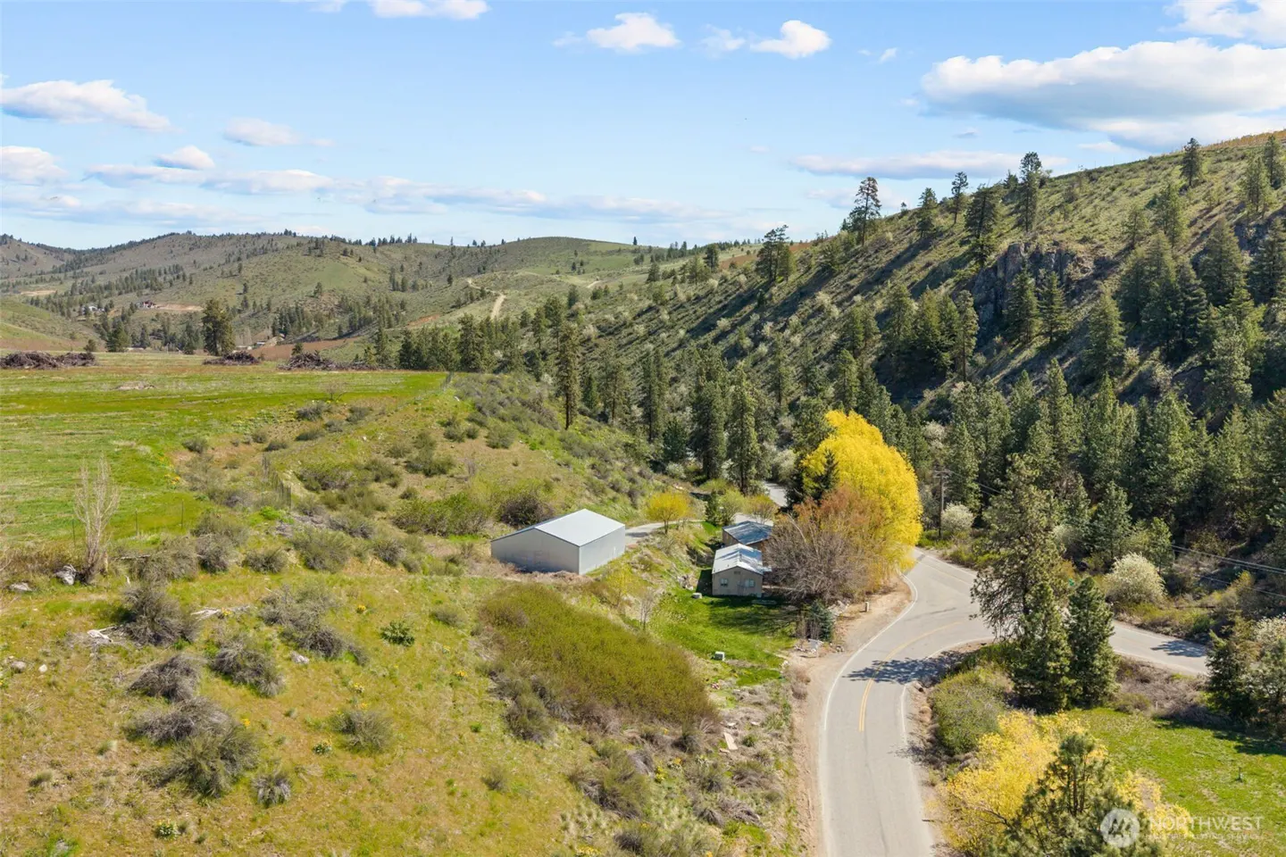 Aerial view of a rural property with a house, barn, and winding road, surrounded by green hills and trees under a blue sky.