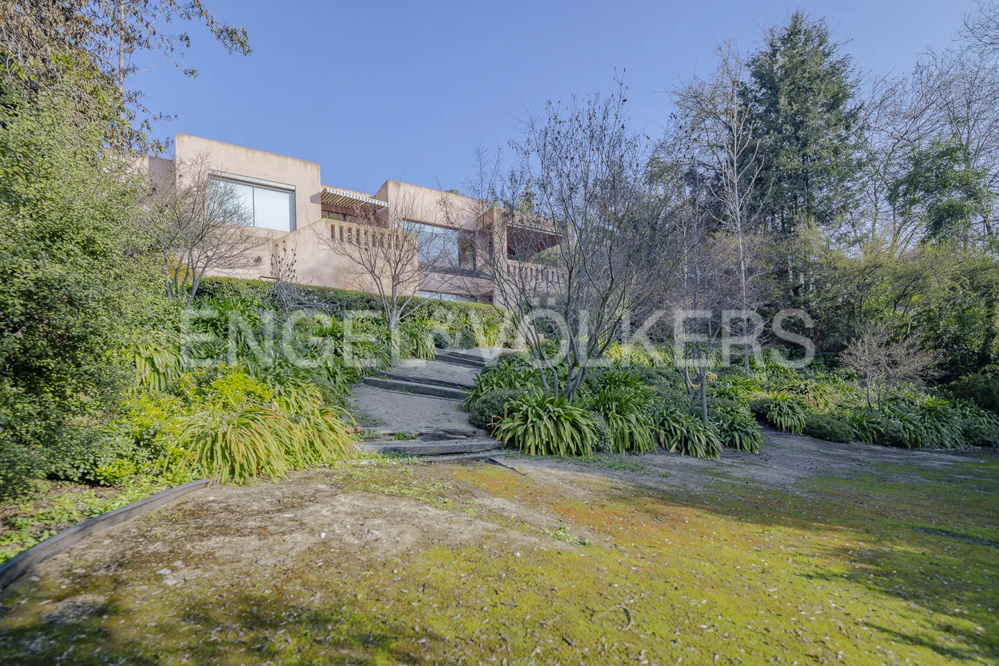 A salmon-colored house sits atop a hill with a stone staircase leading up to it, surrounded by green foliage.