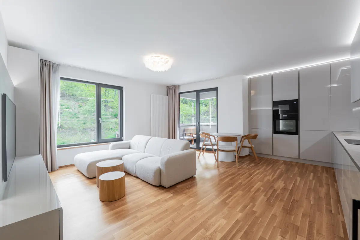 Bright, modern apartment interior with white sofa, wood floors, and kitchen. Large windows overlook green trees.