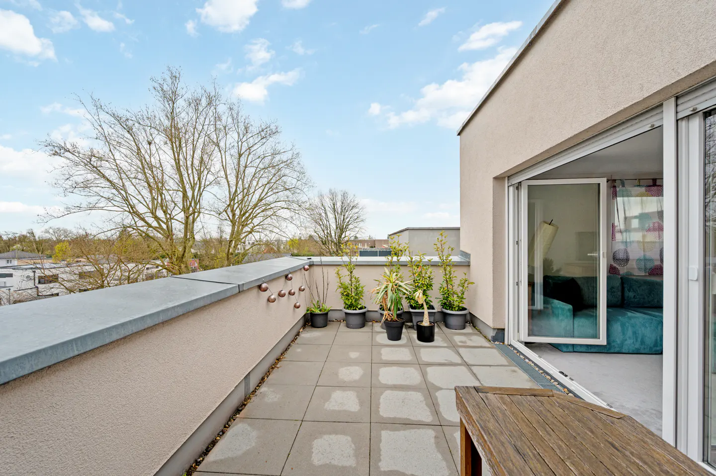 Balcony with gray tiles, potted plants, and a wooden table. Sliding glass doors open to a living room with a teal sofa. Blue sky.