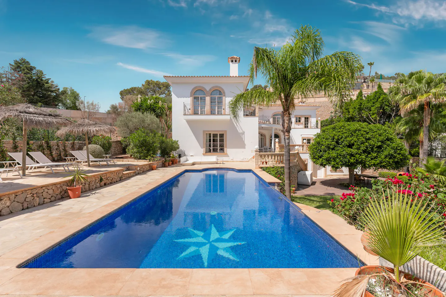 A white two-story house with a blue tiled pool and a star design at the bottom. Palm trees and green foliage surround the house.