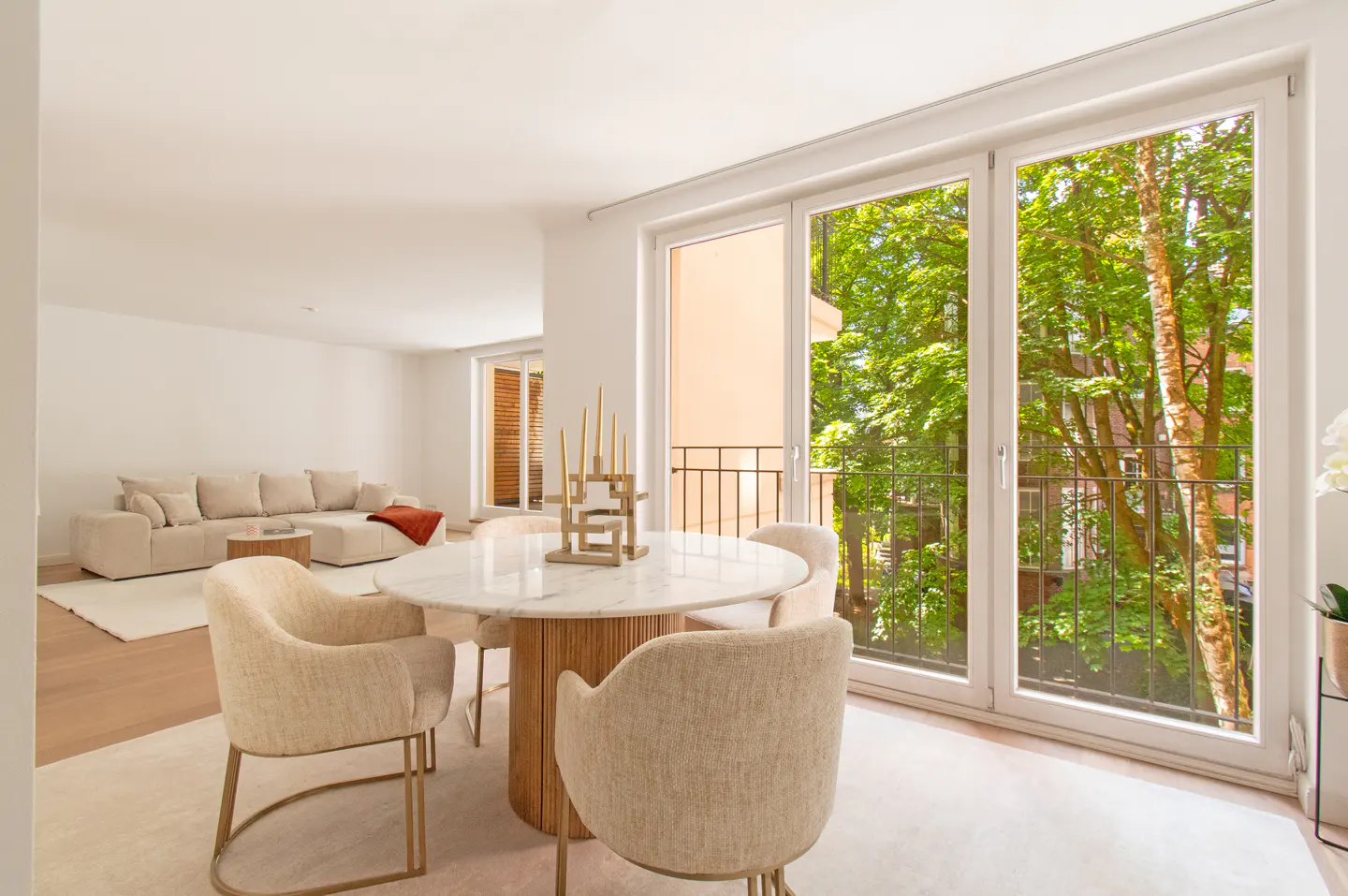 Bright dining area with a marble table, cream chairs, and large glass doors opening to a balcony with lush green trees.
