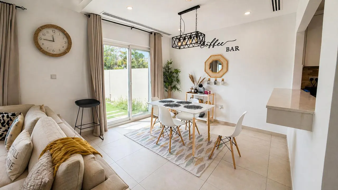 Bright dining area with white table, chairs, and patterned rug. A "Coffee Bar" sign and mirror decorate the wall. Sofa and clock are visible.