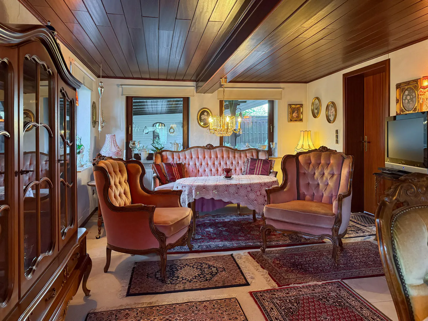 Cozy living room with pink velvet sofa and chairs, dark wood ceiling, and patterned rugs. A large wooden cabinet is on the left.
