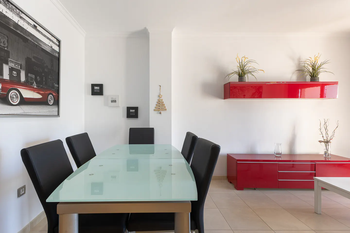 Bright dining room with a glass table, black chairs, and red storage units against white walls. A framed car photo hangs on the wall.