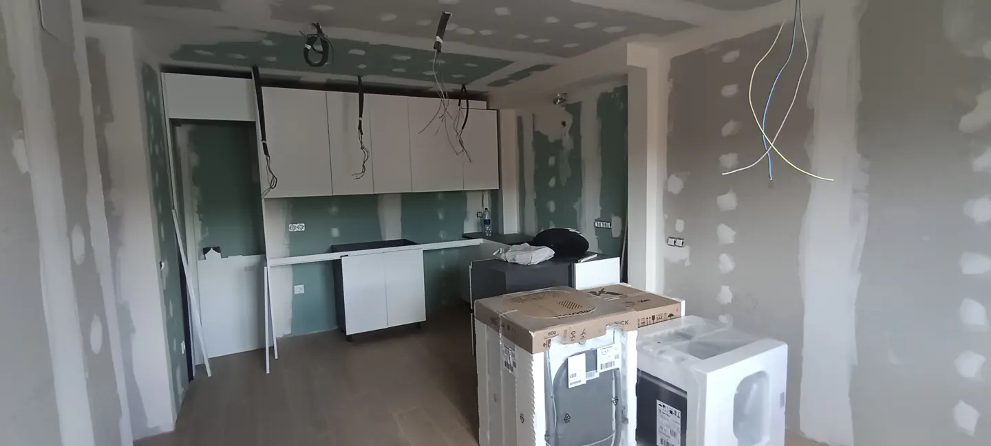 Unfinished kitchen with white cabinets, drywall walls, and exposed wiring. Appliances are in boxes on the floor.
