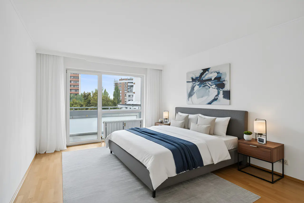 Bright bedroom with a gray bed, white linens, and a blue throw. A balcony door offers a city view. Abstract art hangs above the bed.