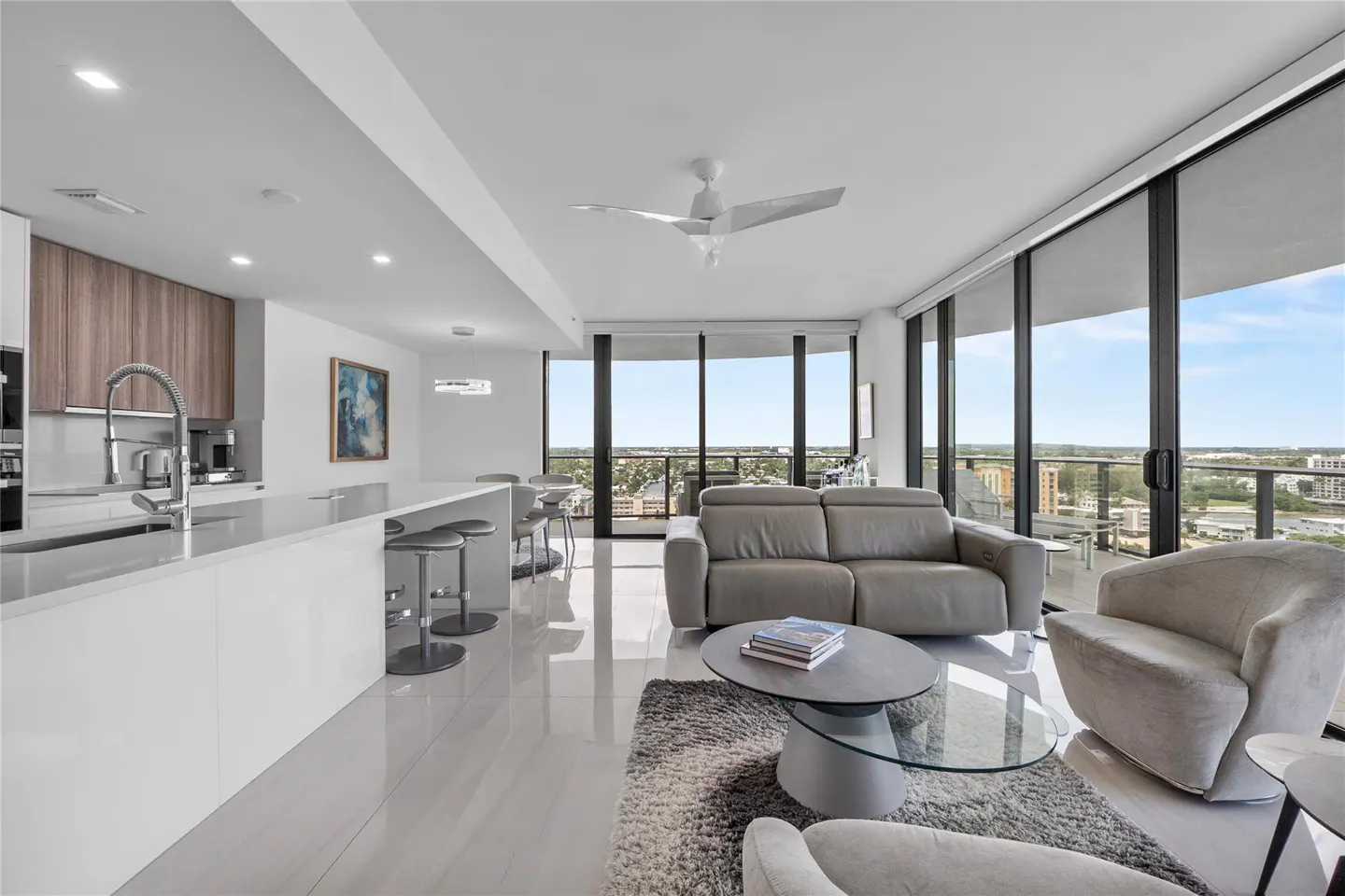 Bright, modern condo interior with white tile floors, gray sofa and chairs, and floor-to-ceiling windows with city views.