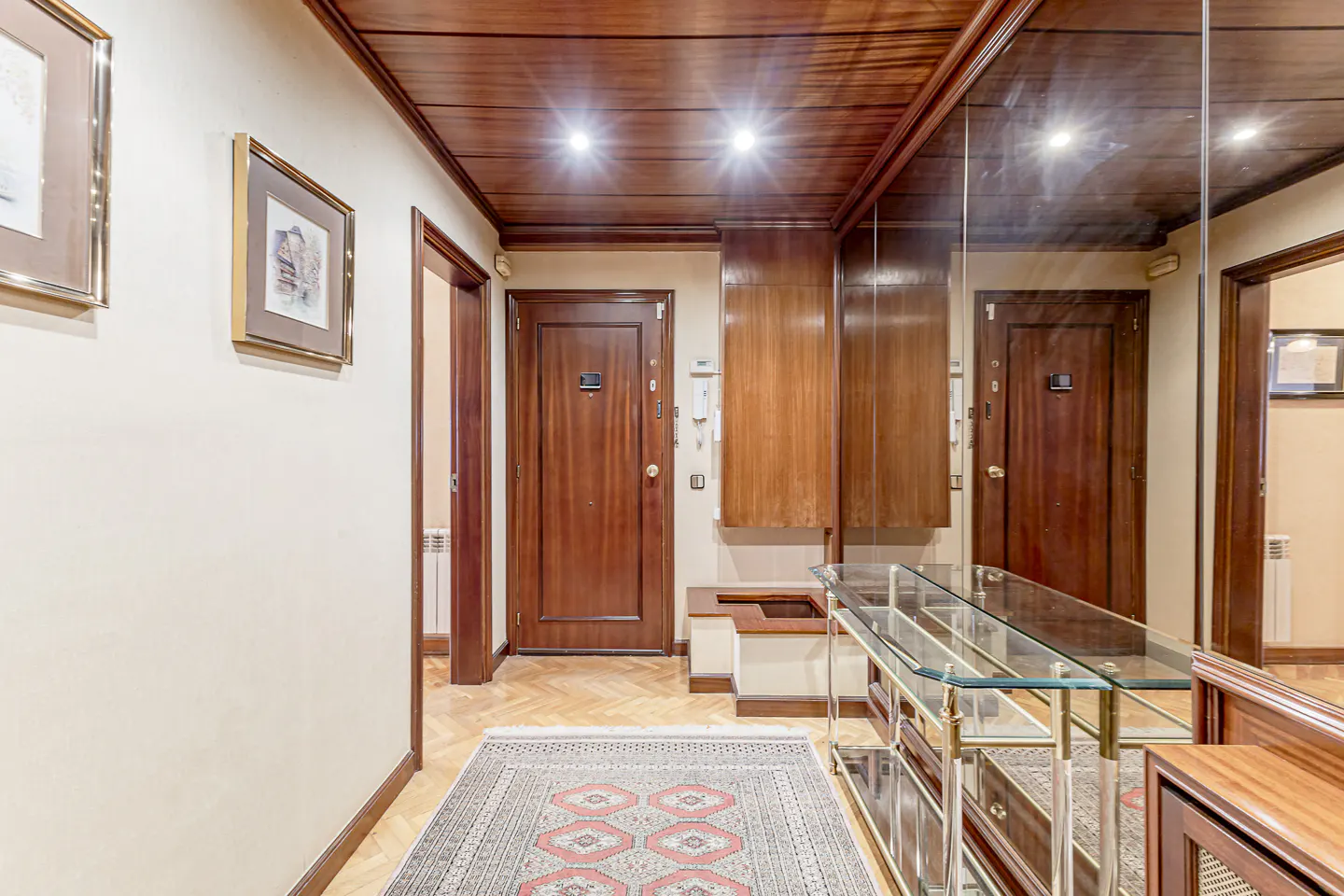Hallway with wood ceiling, parquet floor, and a rug. A glass console table stands near a mirrored wall. Two framed pictures hang on the wall.