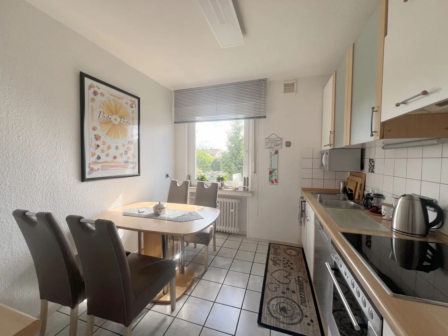 A bright kitchen with a dining table, chairs, and a window with blinds. White cabinets and appliances are visible. A framed pasta poster hangs on the wall.