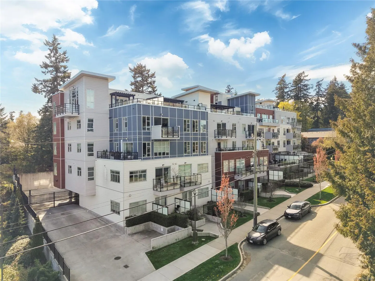 Exterior view of a modern, multi-story apartment building with blue and white facade, balconies, and a gated parking area. Two cars are parked on the street.