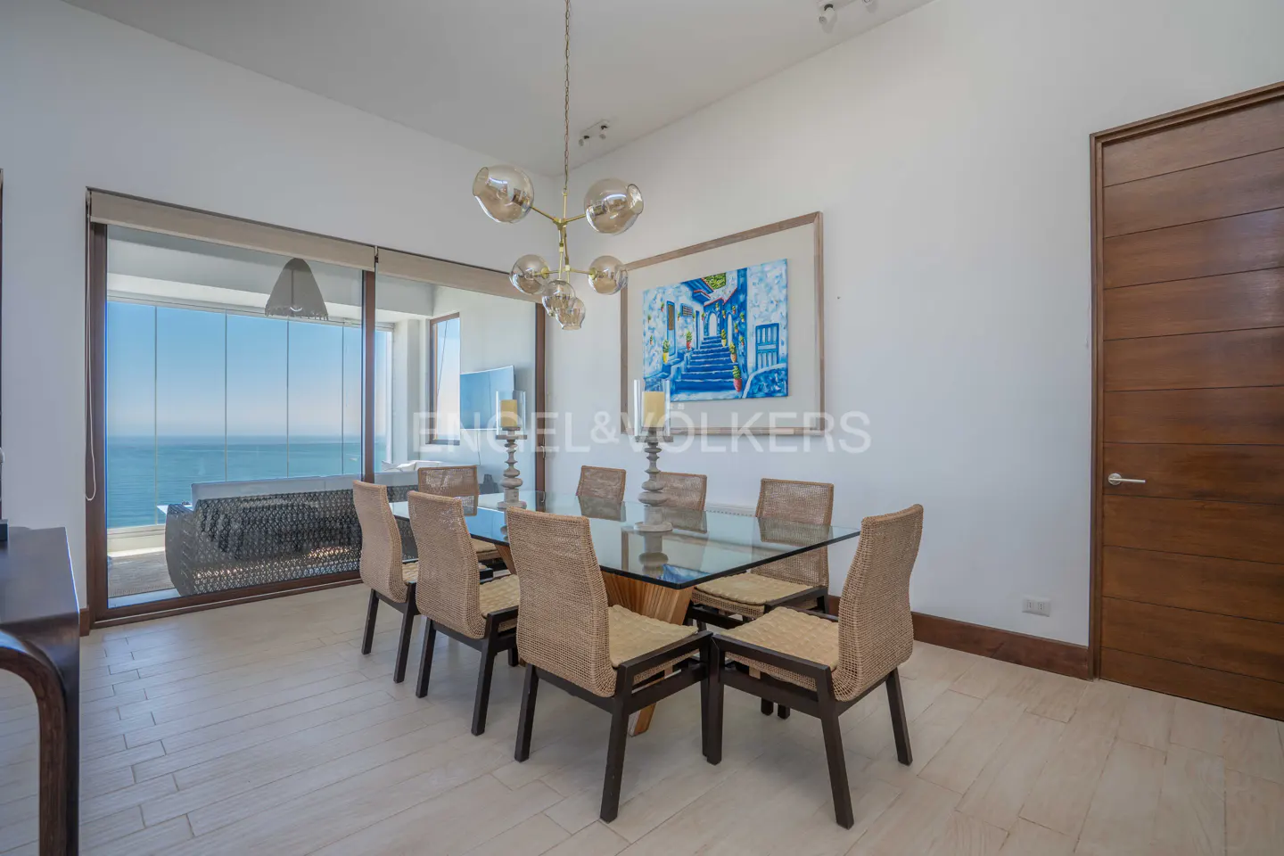 Bright dining room with a glass table, wicker chairs, and ocean view through a large window. A modern chandelier hangs above.