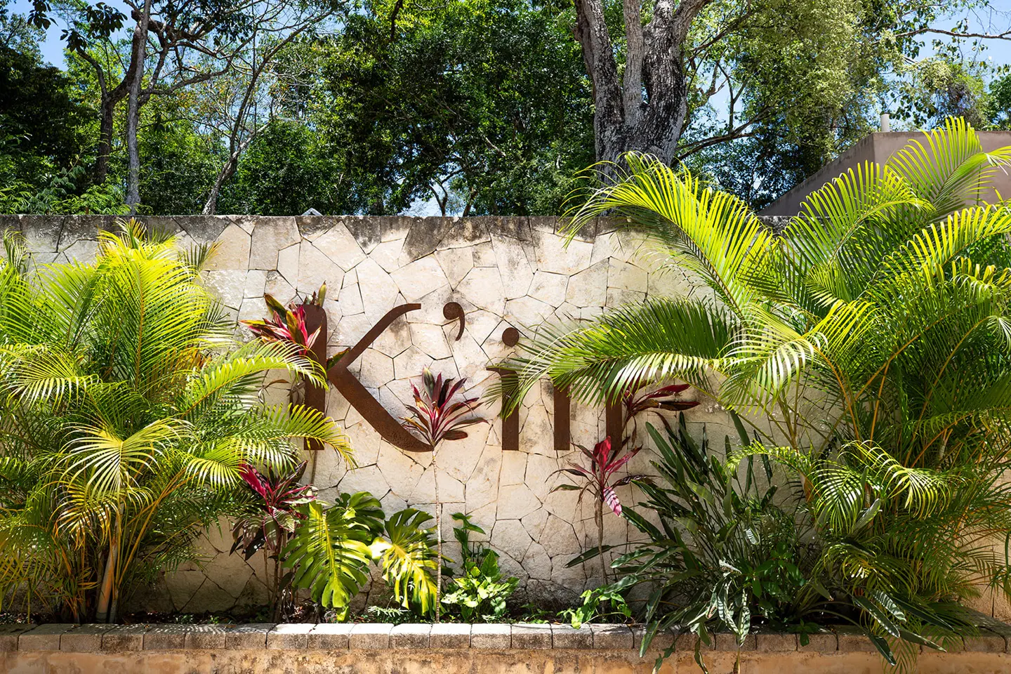 Stone wall with "Kii" metal sign surrounded by lush green tropical plants and trees.