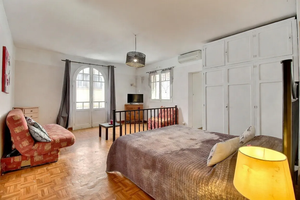 Bedroom with a brown bed, red sofa, and white walls. A black metal railing separates the bed from a TV and chair. A large white wardrobe is on the right.