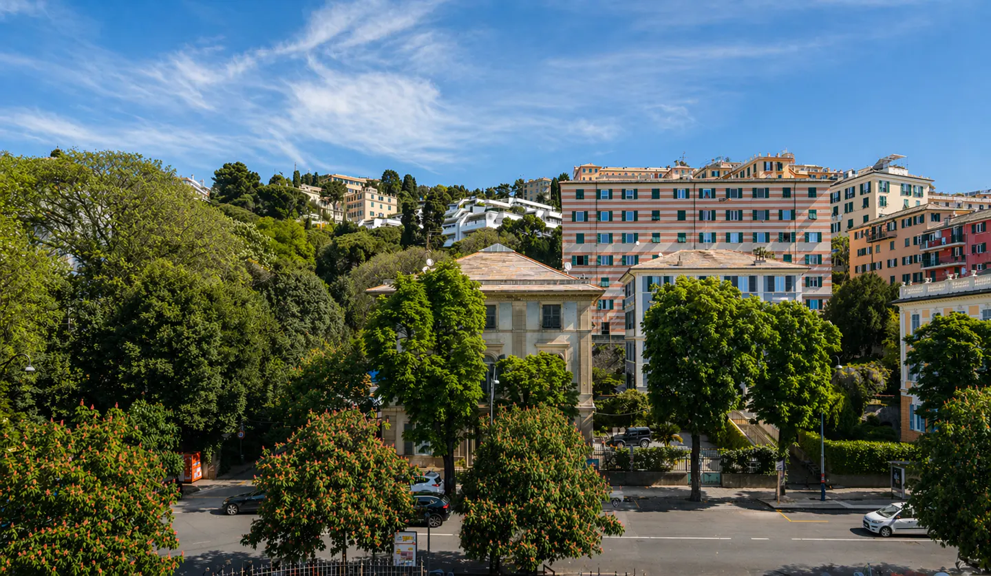 Cityscape view of Genoa, Italy, featuring buildings, trees, and a blue sky with wispy clouds. A street with cars runs in front.