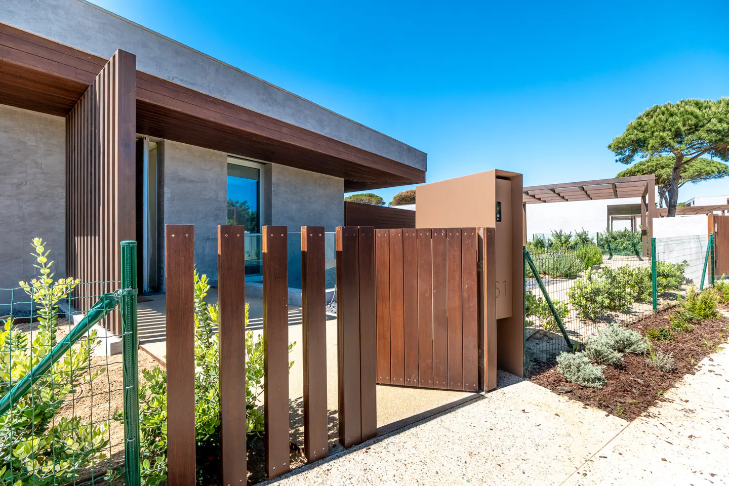 Modern home exterior with a brown wooden gate, gray stucco walls, and a blue sky background. Landscaped garden and walkway visible.