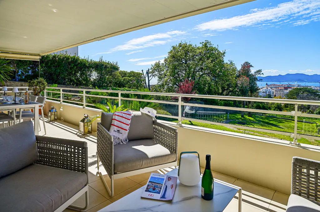 Balcony with gray woven chairs, white table, champagne, and a view of trees, city, and ocean under a blue sky.