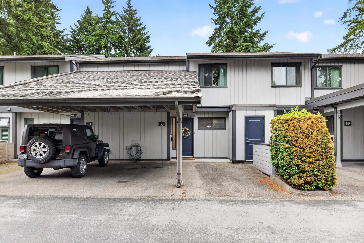 Grey townhouse exterior with a covered carport, a black Jeep parked inside, and a large green bush in the front yard.