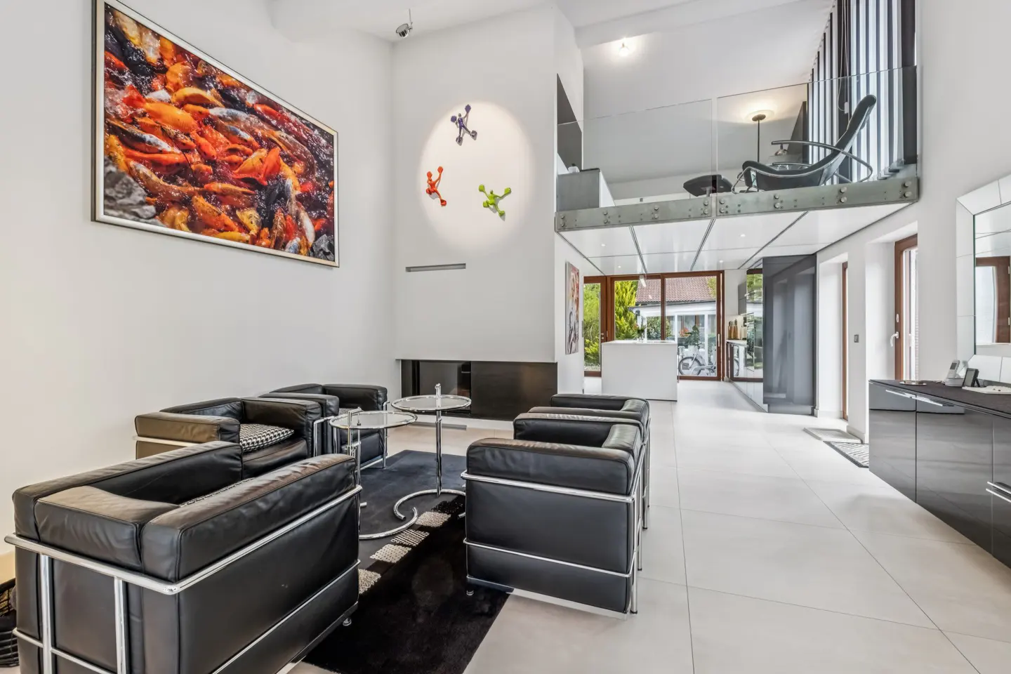 Bright, modern living room with black leather chairs, glass tables, and a large koi fish painting. A loft area overlooks the space.