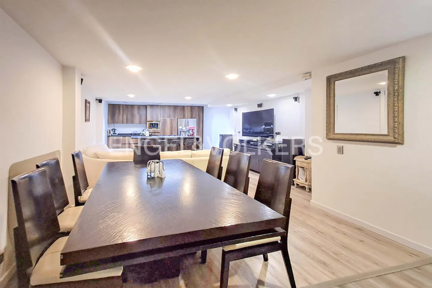 Open-concept living space with a dark wood dining table and chairs, leading to a kitchen and living room with a TV.