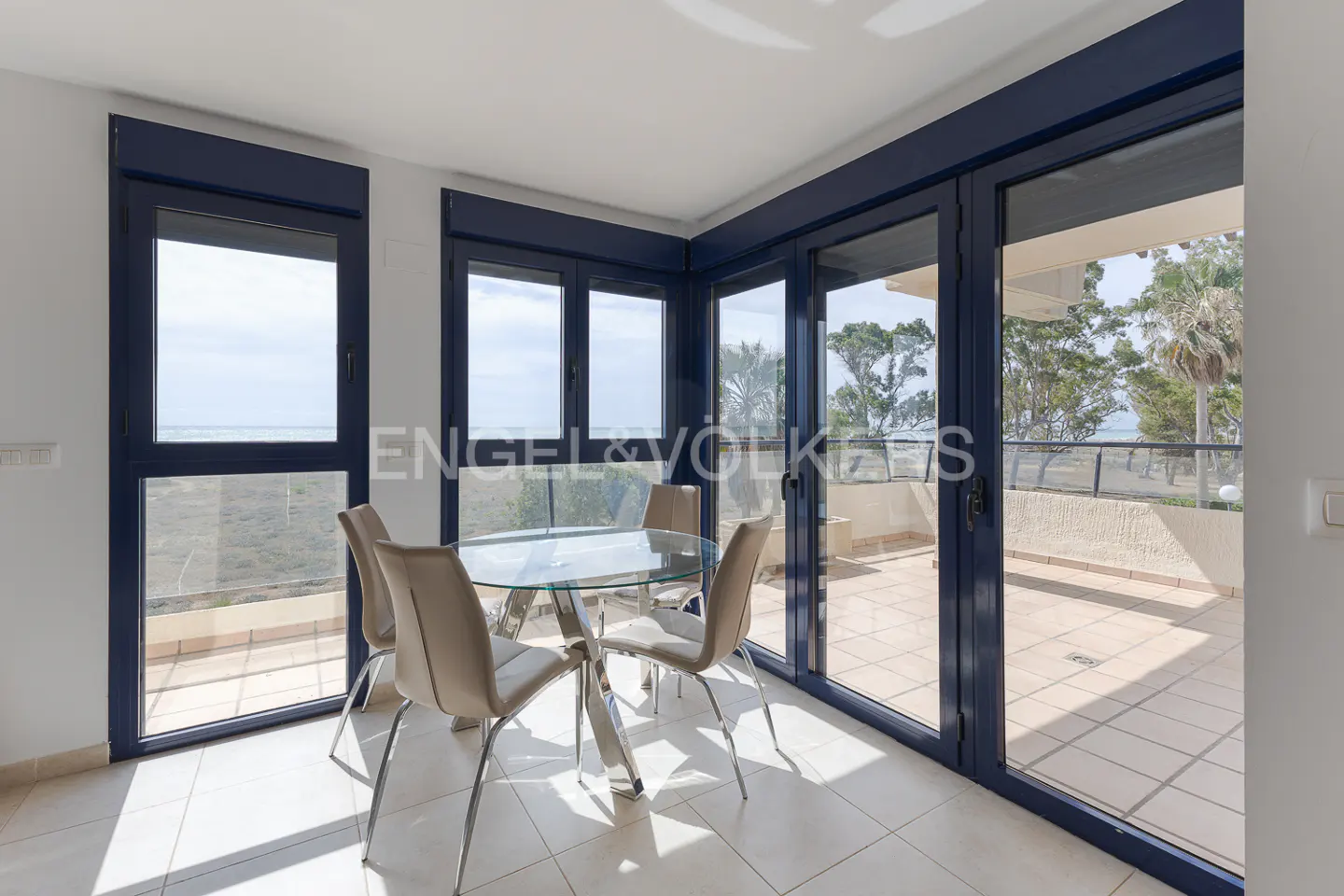 Dining area with a glass table and four chairs, blue window frames, and a view of the ocean.