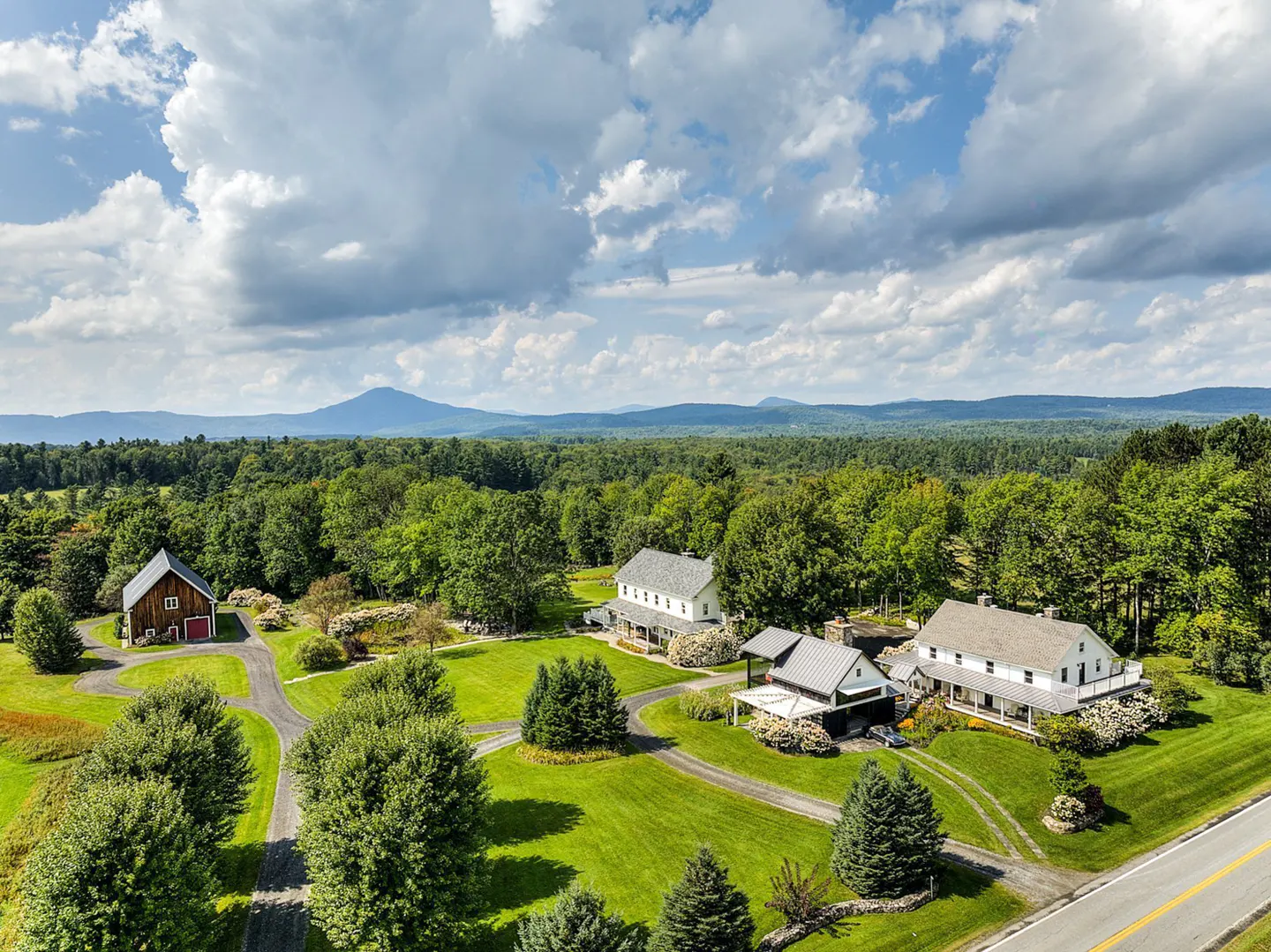 Aerial view of a rural estate with white houses, a red barn, green lawns, trees, and mountains in the background under a cloudy sky.