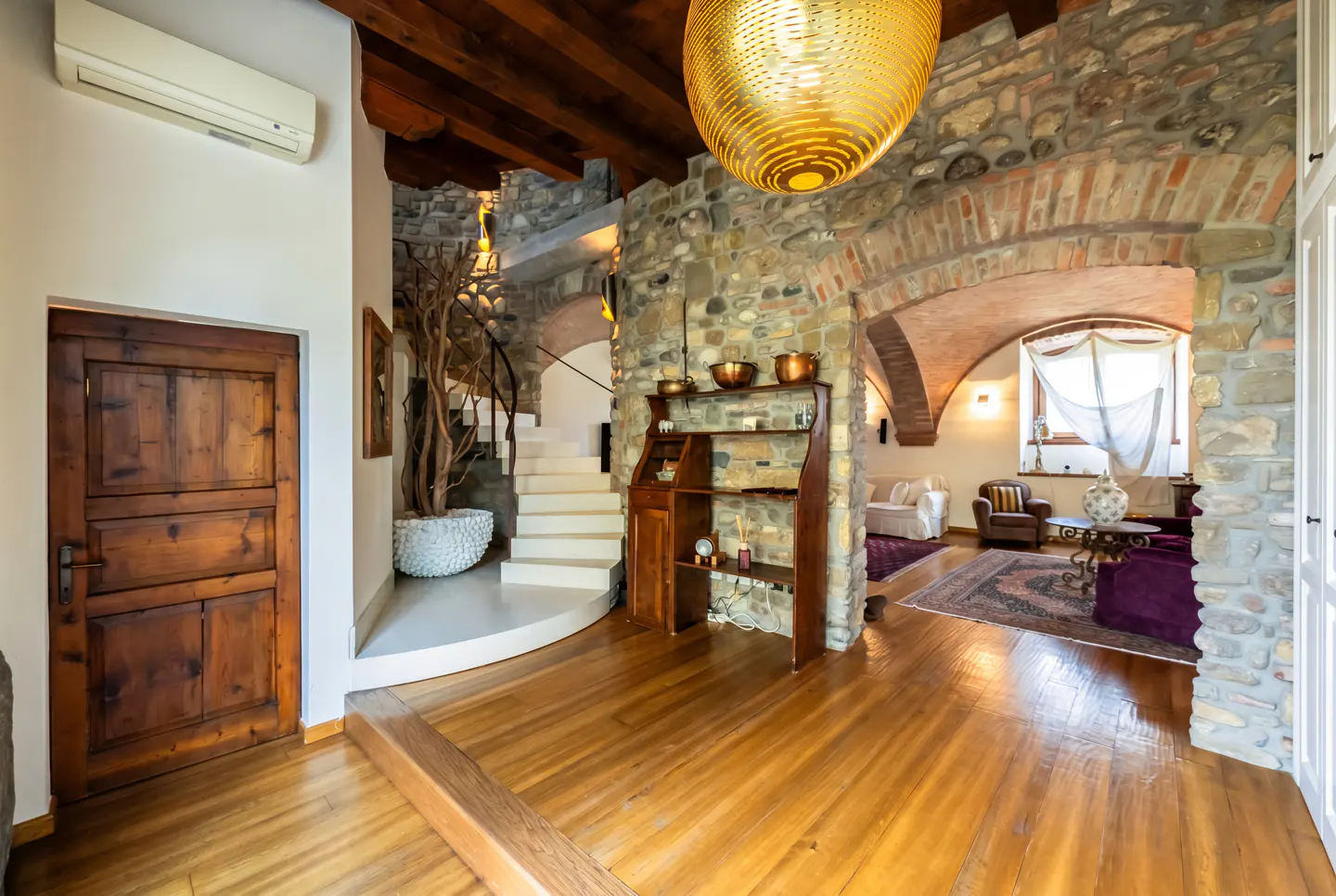 Interior view of a home with wood floors, stone walls, and arched doorways. A staircase and living room are visible.