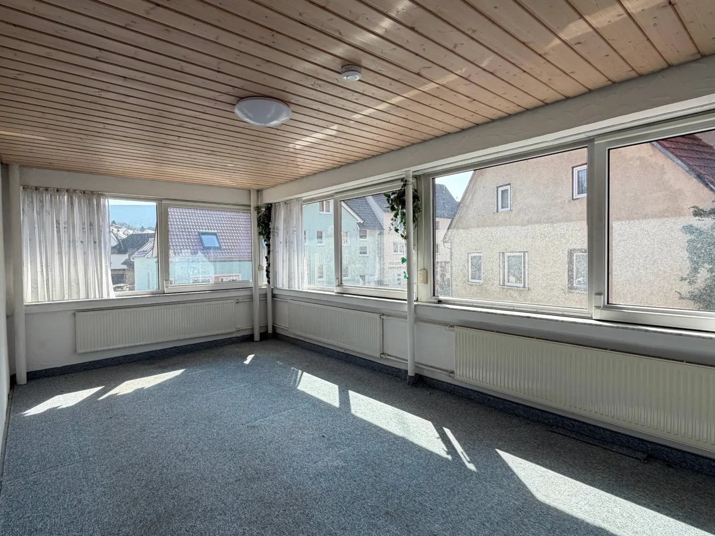 Bright sunroom with wood paneled ceiling, blue carpet, and white walls. Large windows offer views of houses and sky. White curtains hang on the left.