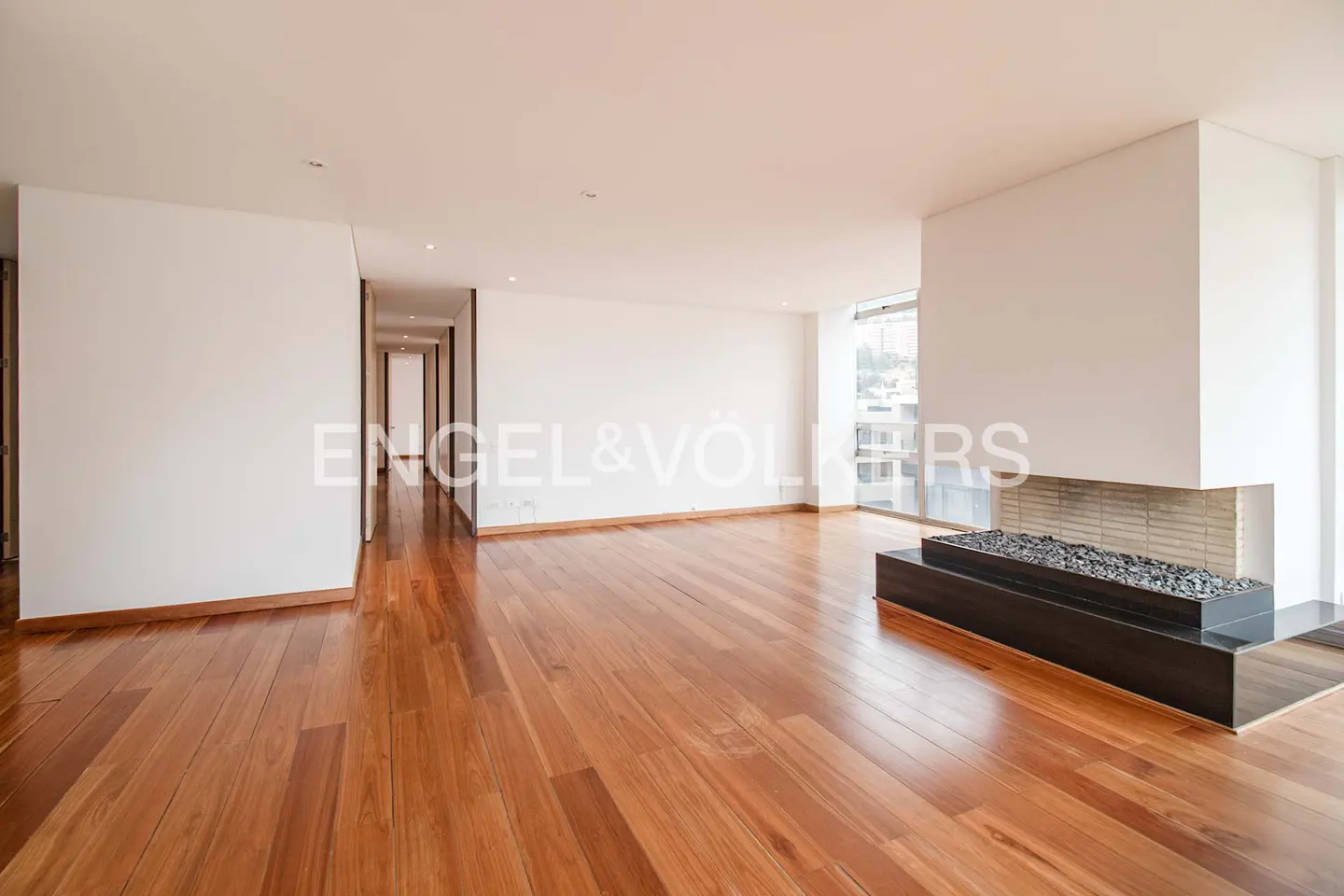 Bright, empty living room with hardwood floors, white walls, and a modern fireplace with black stones. A hallway is visible in the background.