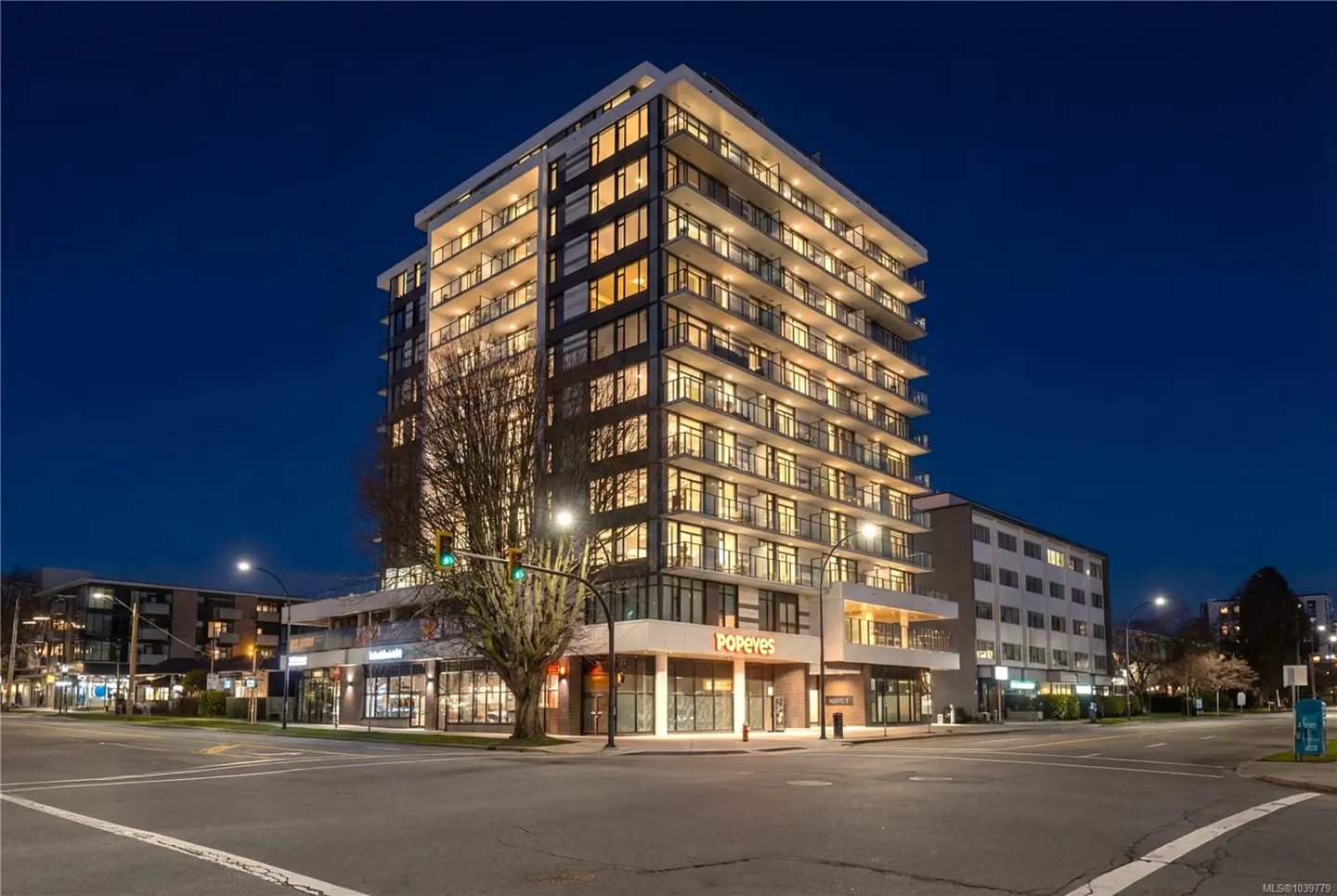 A modern high-rise apartment building at dusk with a Popeyes restaurant on the ground floor, viewed from a street corner.
