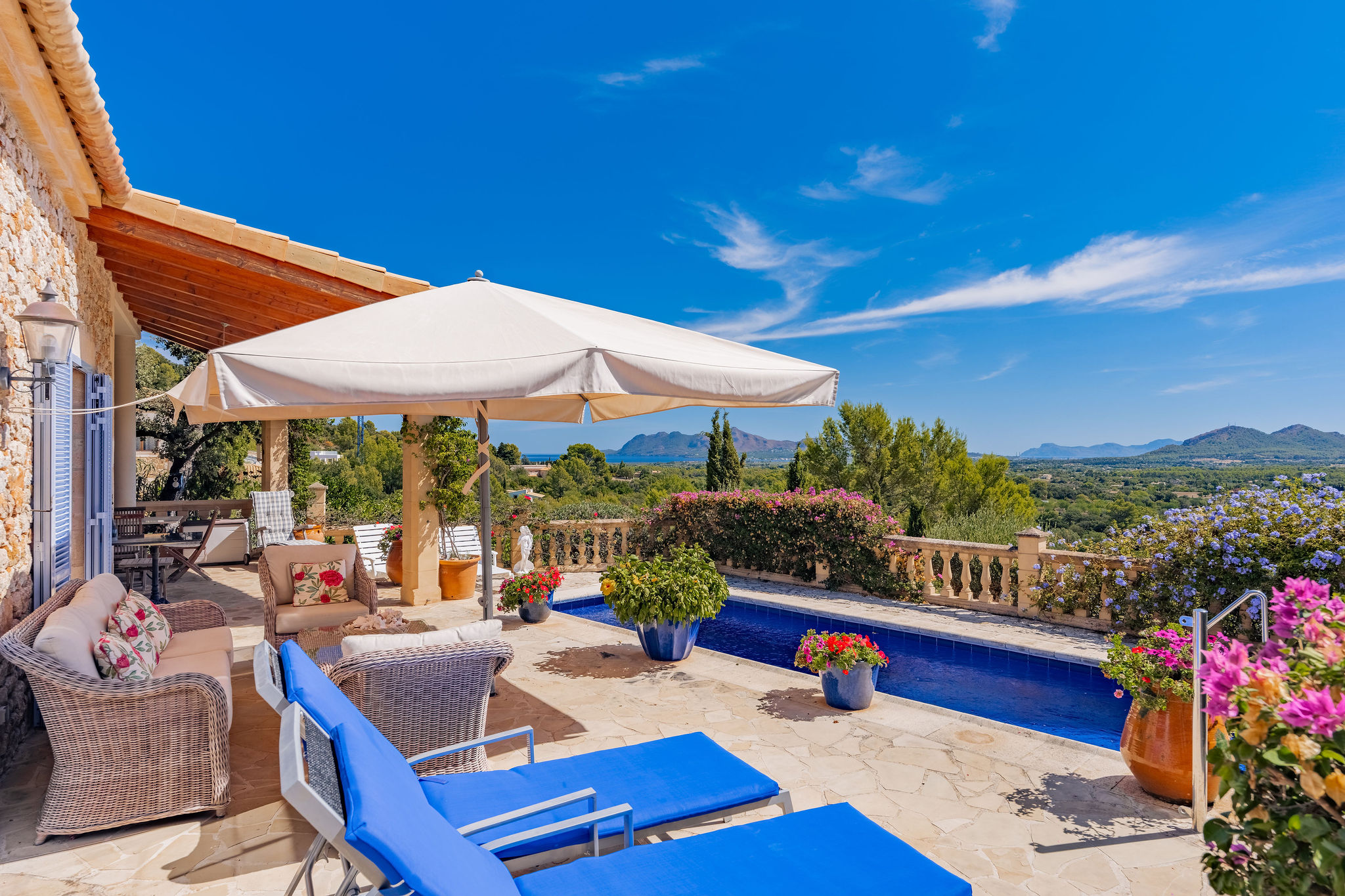Stone patio with a pool, blue lounge chairs, white umbrella, and potted flowers overlooking a scenic mountain landscape. [AI-generated text]
