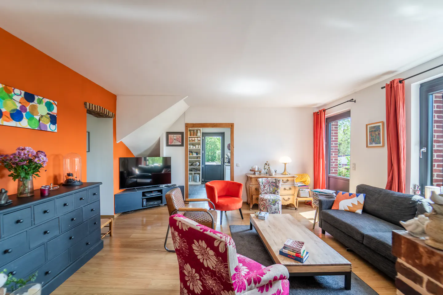 Bright living room with hardwood floors, orange accent wall, gray sofa, and floral armchairs. A TV and dresser are on the left.