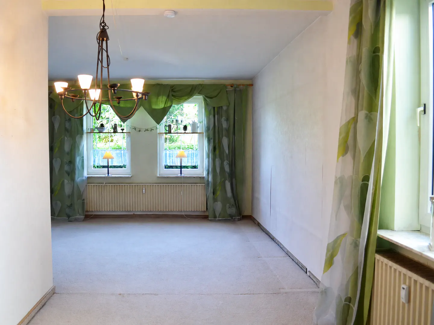 Empty room with white walls, beige carpet, a rustic chandelier, and windows draped in green leaf-patterned curtains.