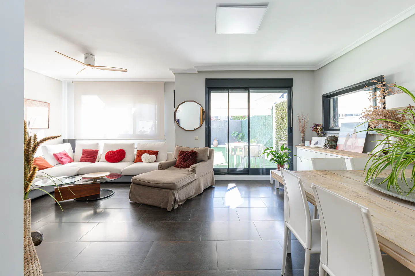 Bright living room with white sofa, chaise lounge, and dining table. Black sliding doors lead to a green backyard.