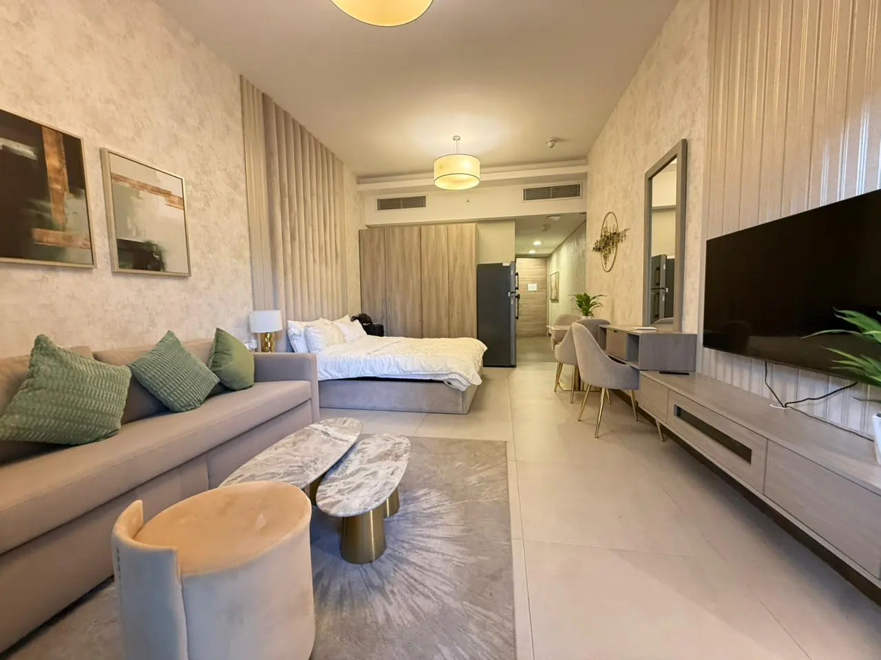 A modern studio apartment with a sofa, bed, TV, dining table, and kitchen appliances. The color scheme is neutral with green accents.