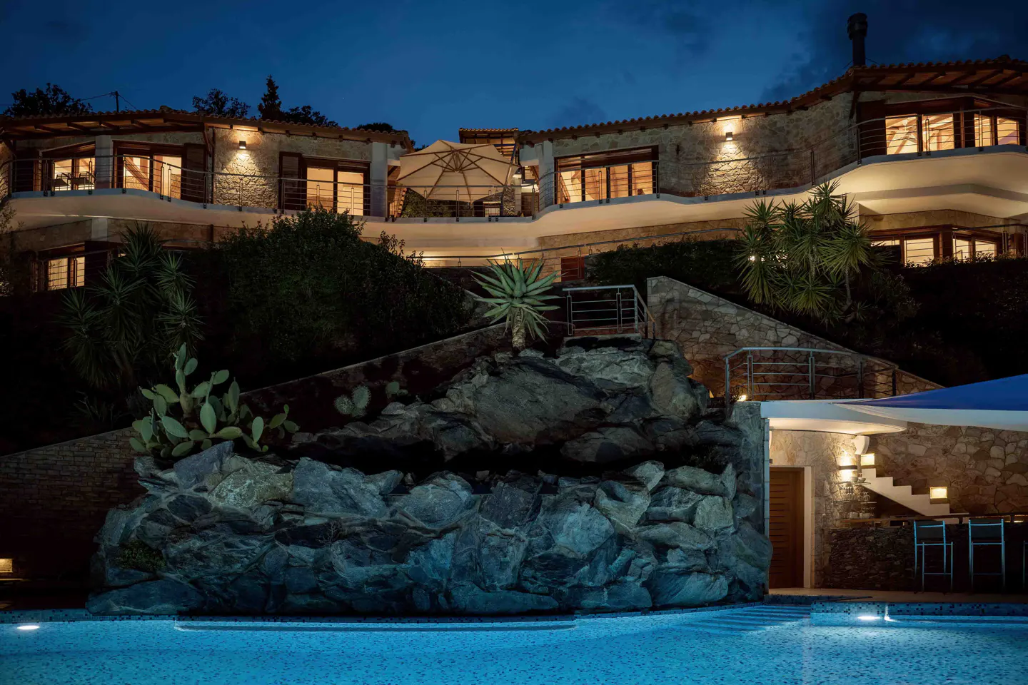 Night view of a stone house with lit windows, a pool, and rock features.