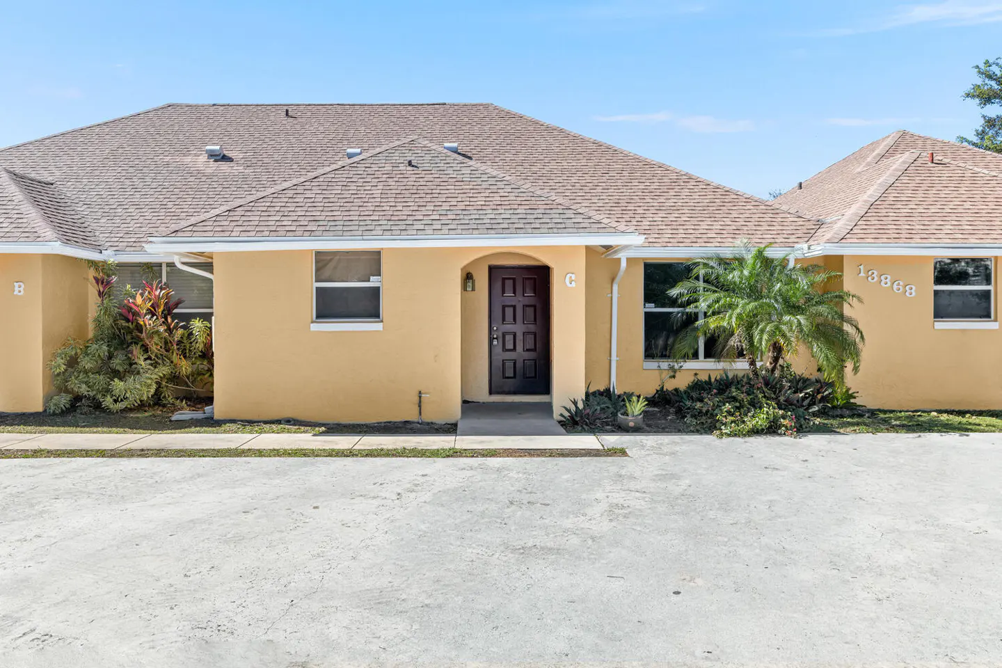 Tan duplex with a brown shingle roof, dark brown front door, and a small palm tree in front. Blue sky above.
