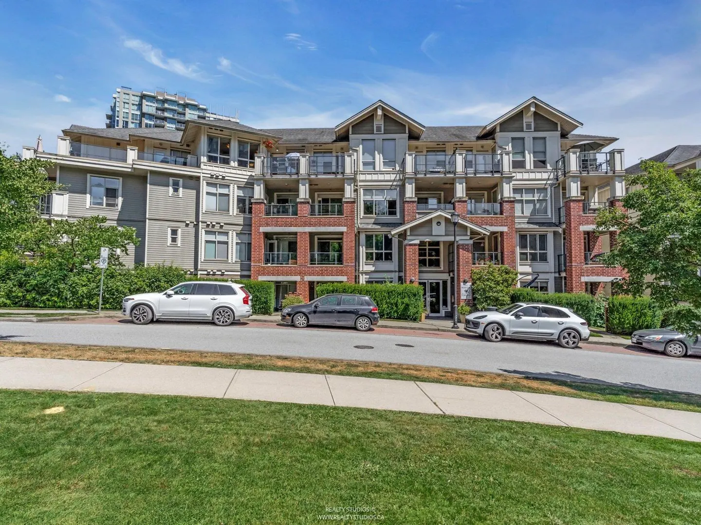 Exterior view of a three-story apartment building with gray siding and brick accents, cars parked on the street.