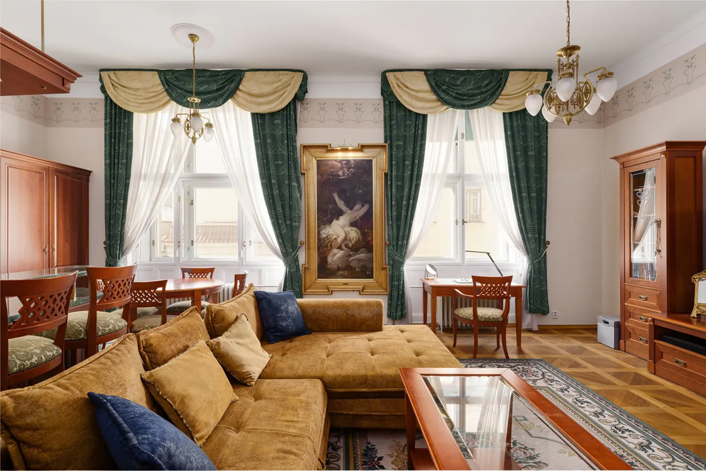 Living room with a brown sectional sofa, blue pillows, and a glass coffee table. Windows have green and gold curtains. A painting hangs between the windows.
