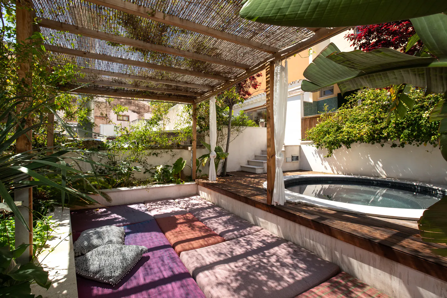 Outdoor patio with a wooden pergola, colorful cushions, and a small round pool. Green plants and white walls surround the space.