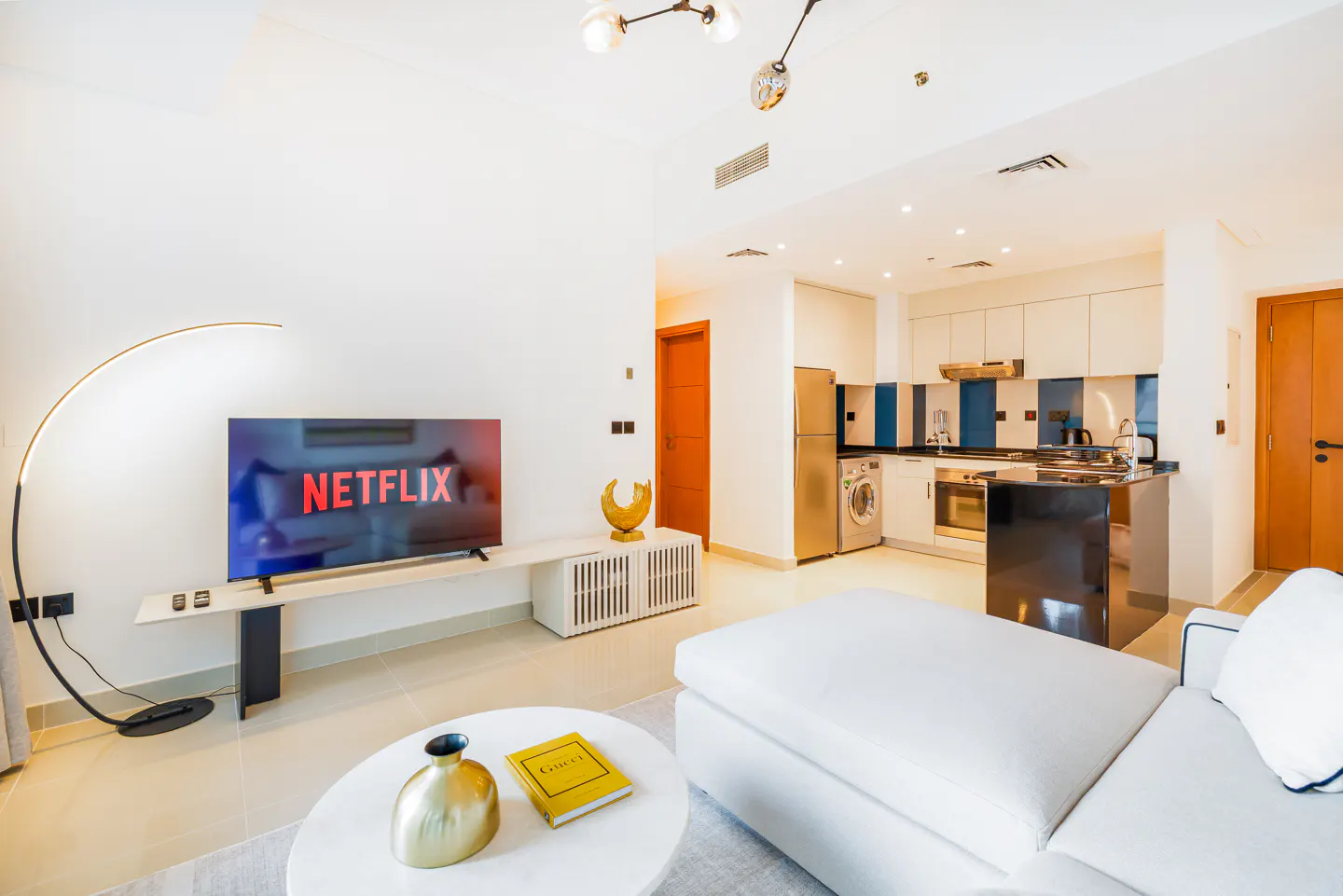 Bright, modern apartment interior with white walls, a white sectional sofa, and a TV displaying Netflix. A kitchen area is visible in the background.