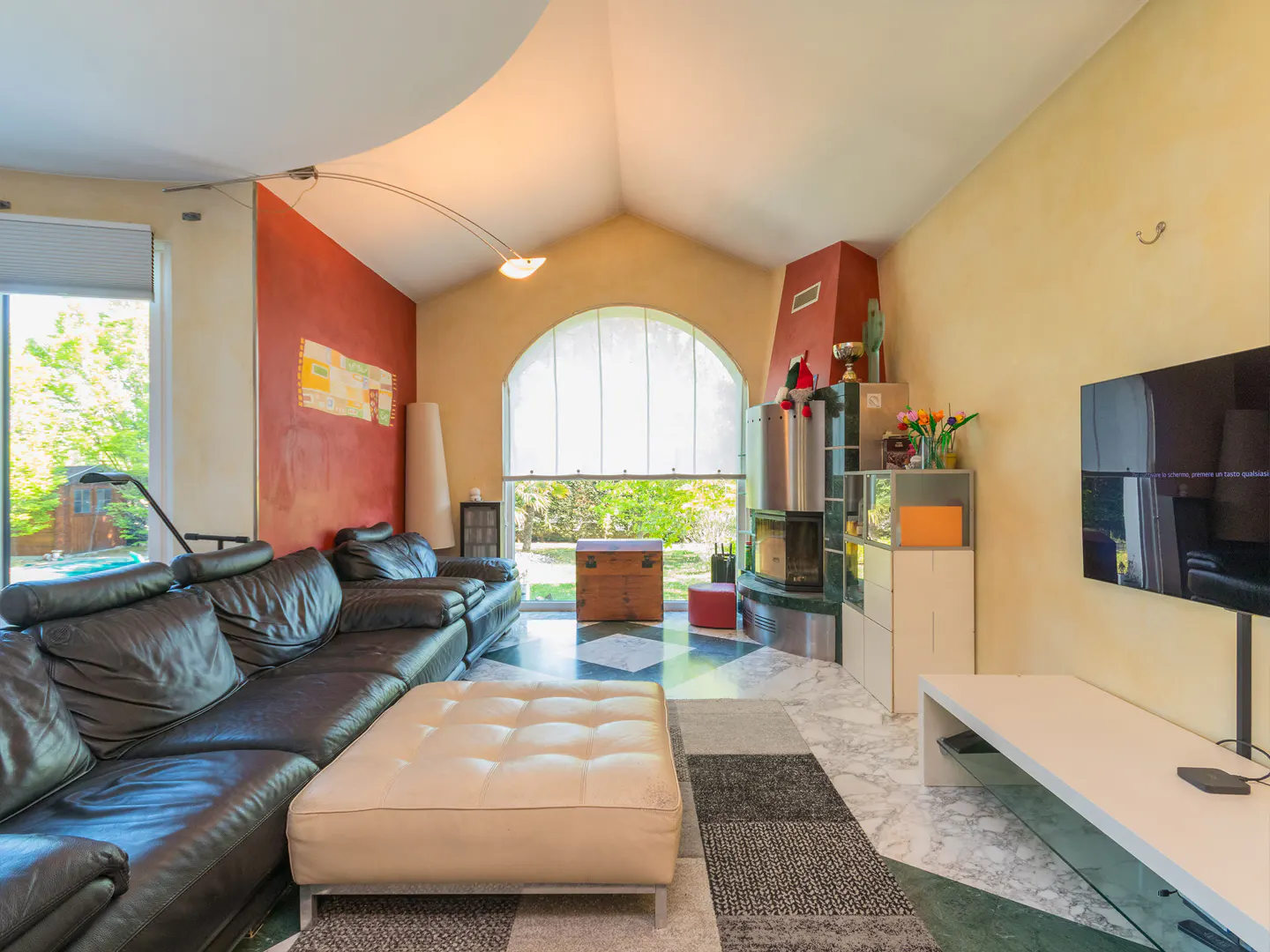 Living room with black leather sofa, cream ottoman, fireplace, and TV. Walls are yellow and red. Large window with view of greenery.