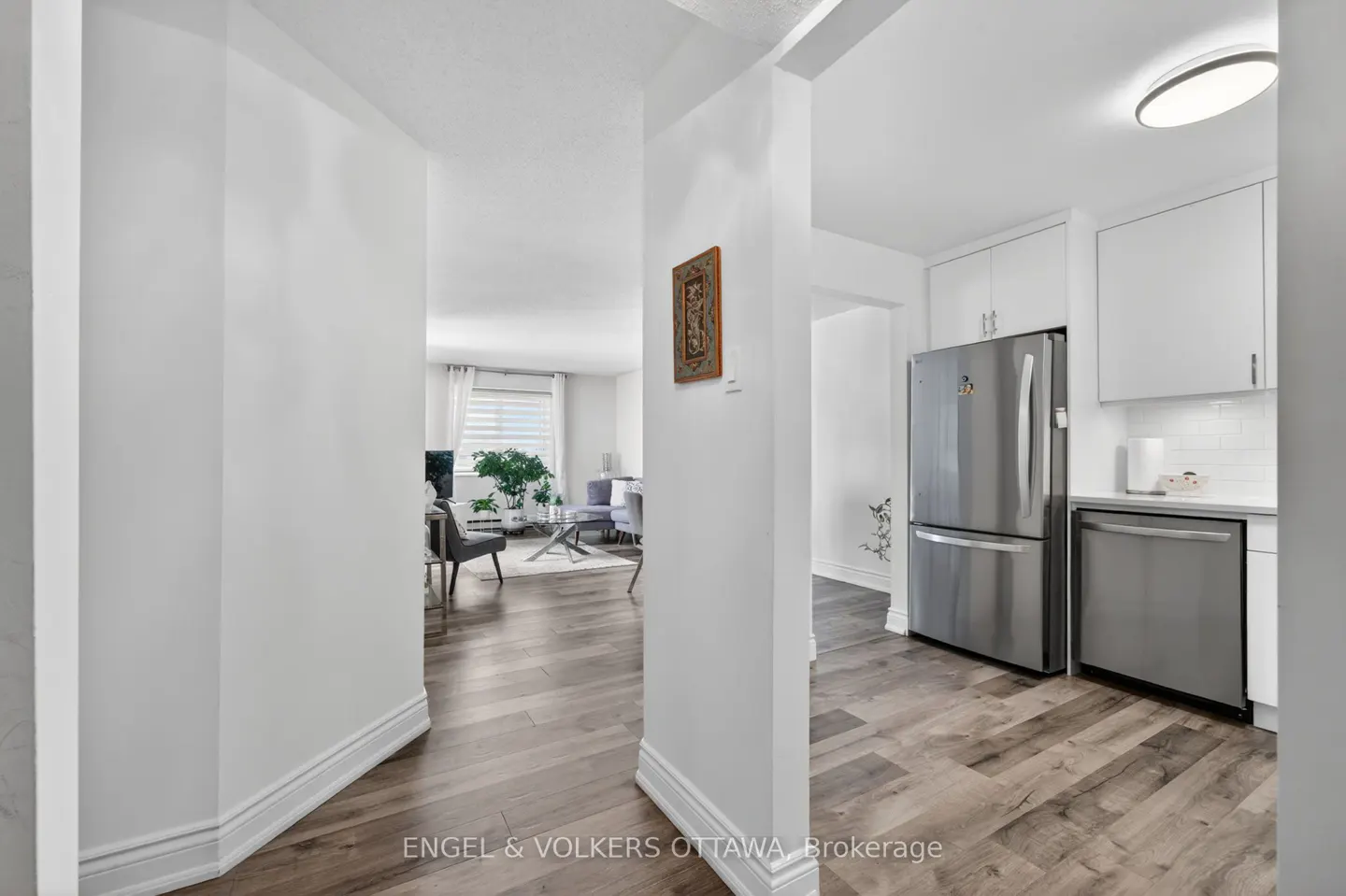A bright condo interior with hardwood floors, white walls, and stainless steel appliances. A living room is visible in the background.
