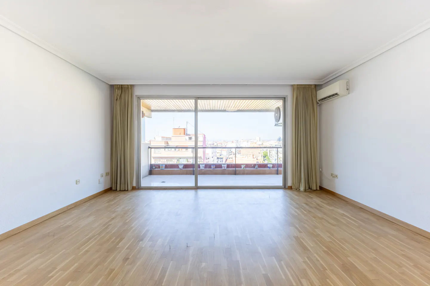 Bright, empty room with wood floors, white walls, and large sliding glass doors to a balcony overlooking a city.