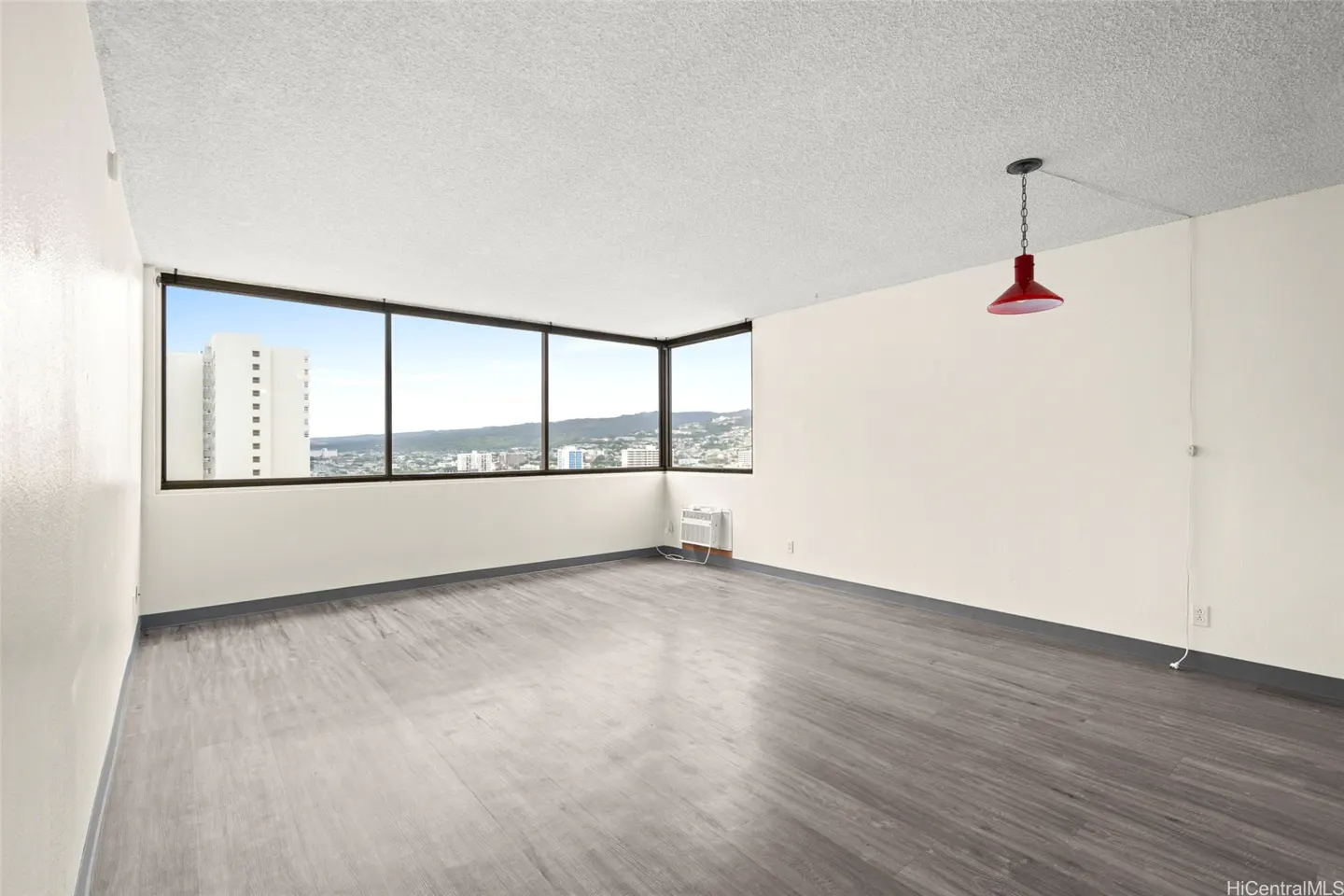 Empty room with gray wood floors, white walls, and a large window with a city view. A red pendant light hangs from the ceiling.