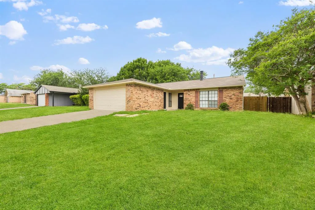 A single-story brick house with a green lawn under a blue sky with white clouds. A driveway leads to an attached garage.
