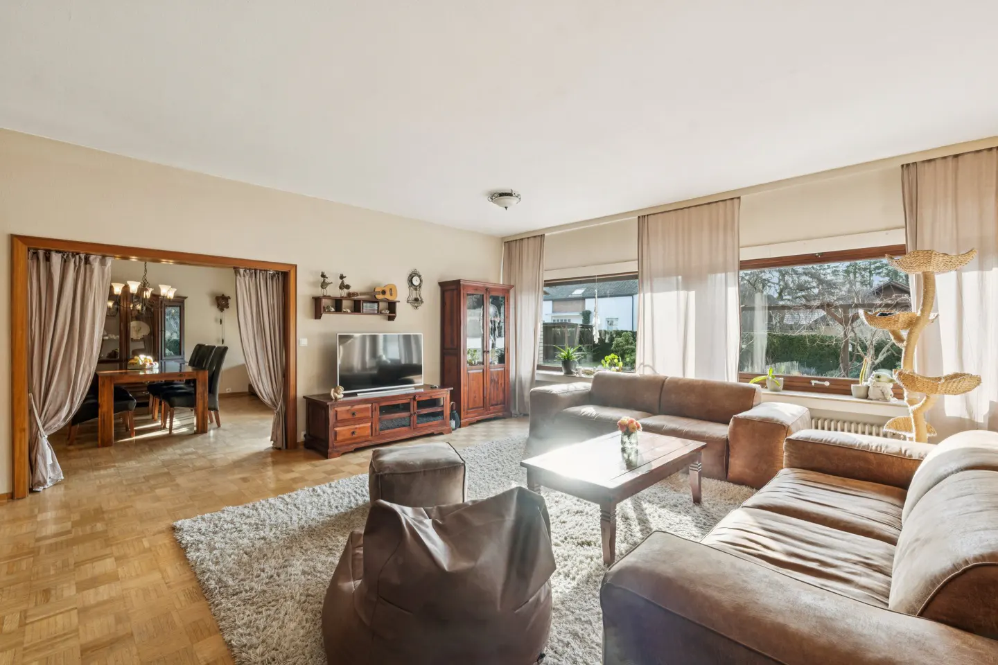 Bright living room with brown sofas, wood floors, and a dining room visible through a curtained doorway. A TV and cat tree are also present.